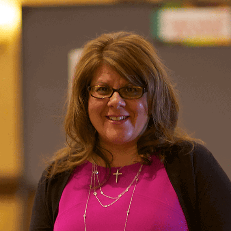 A smiling woman with glasses and shoulder-length hair at the SIIM Annual Meeting, wearing a pink top and layered necklaces.