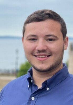 A smiling young man in a blue shirt outdoors near water, attending the SIIM Annual Meeting 2023, showcasing professional engagement in medical imaging and informatics.
