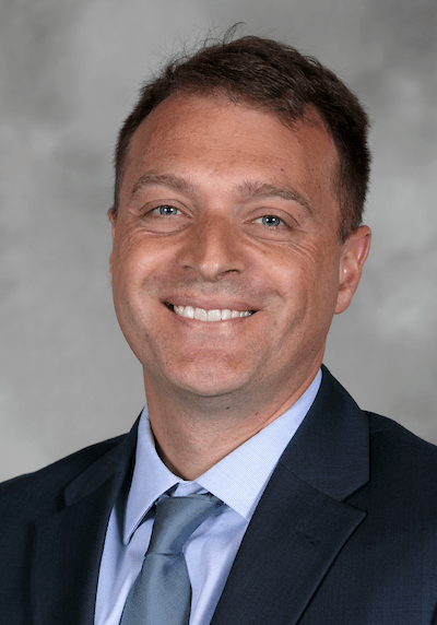 1. Professional male at SIIM Annual Meeting wearing a suit, smiling, with a gray background; radiology imaging, medical innovation, healthcare technology.