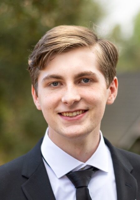 Young male medical professional smiling at SIIM Annual Meeting, wearing formal suit, with outdoor background.