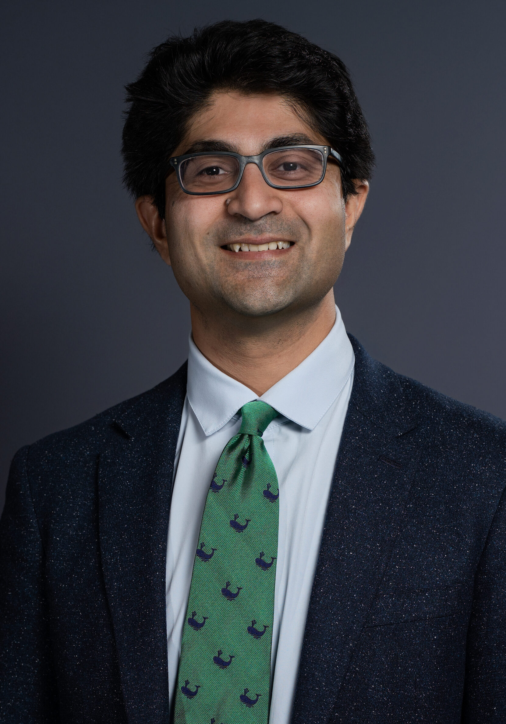 A professional portrait of a smiling man wearing glasses, a dark blazer, white shirt, and a green tie with navy blue patterns, set against a plain dark background.