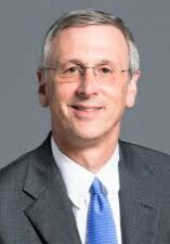 A person in a gray suit and blue tie smiling at the camera against a plain gray background.