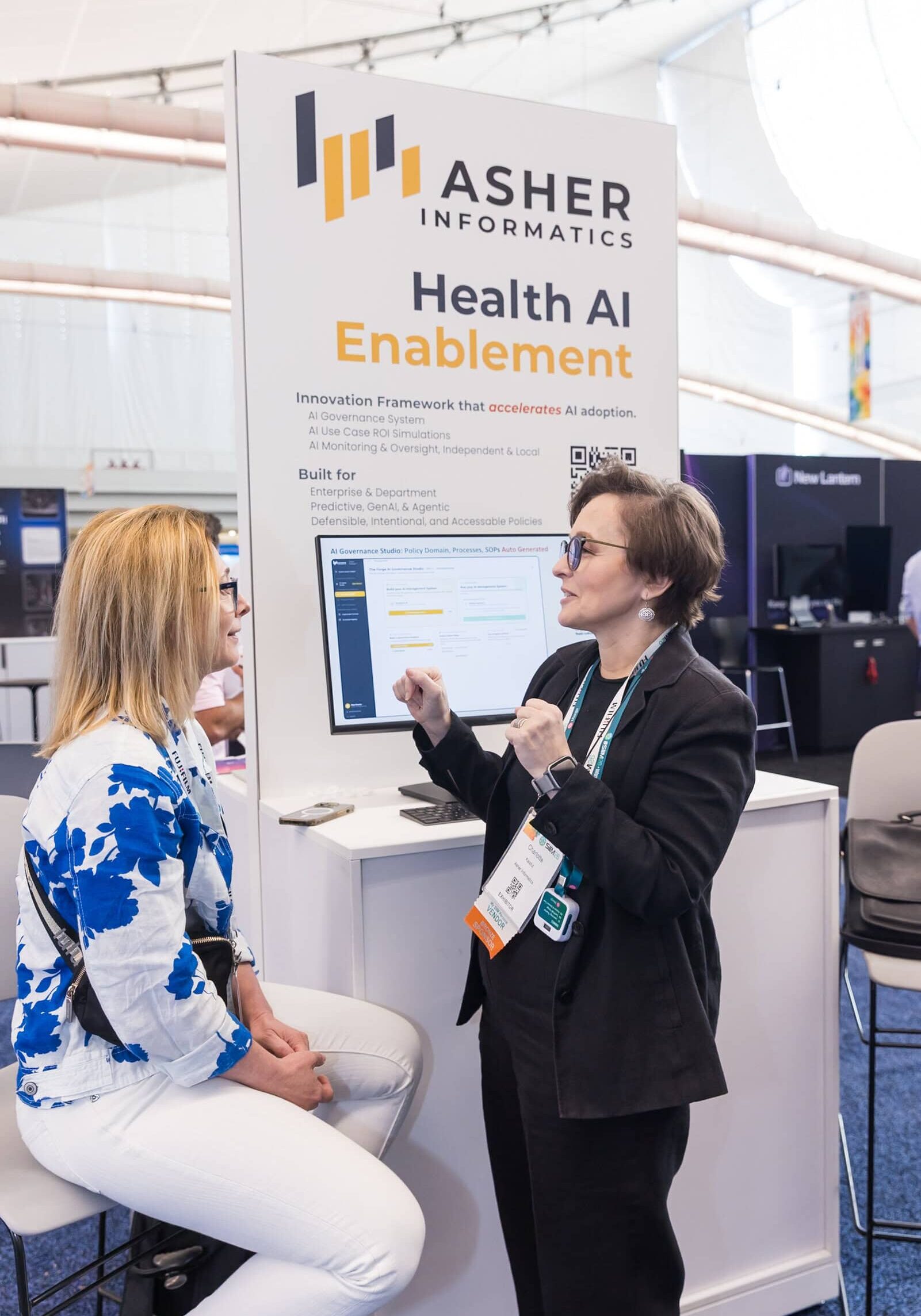 Two women engaged in a discussion at the SIIM Annual Meeting booth showcasing ASHER Informatics' Health AI Enablement platform.