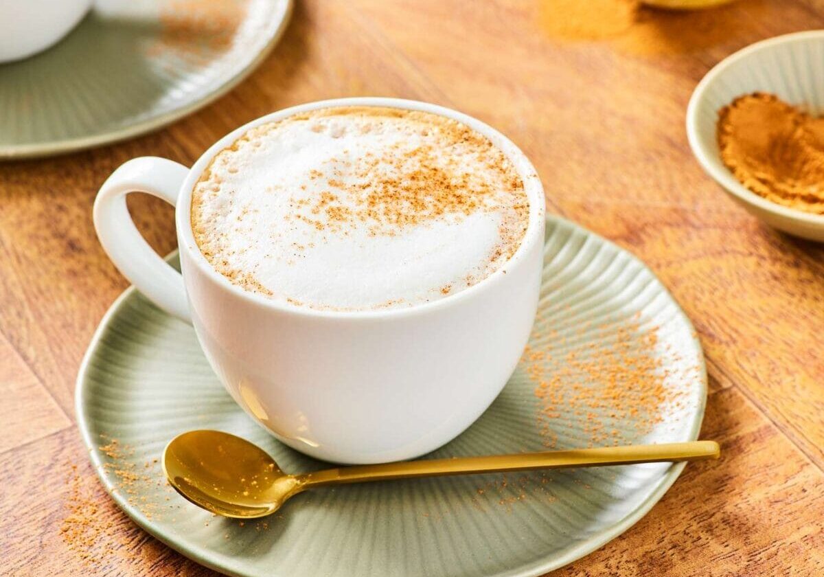 Coffee with whipped cream and cinnamon on a ceramic saucer with a golden spoon, served on a wooden table.
