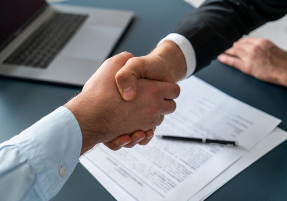 Close-up of two professionals shaking hands over a business agreement at the SIIM Annual Meeting, with documents and a laptop on the table.
